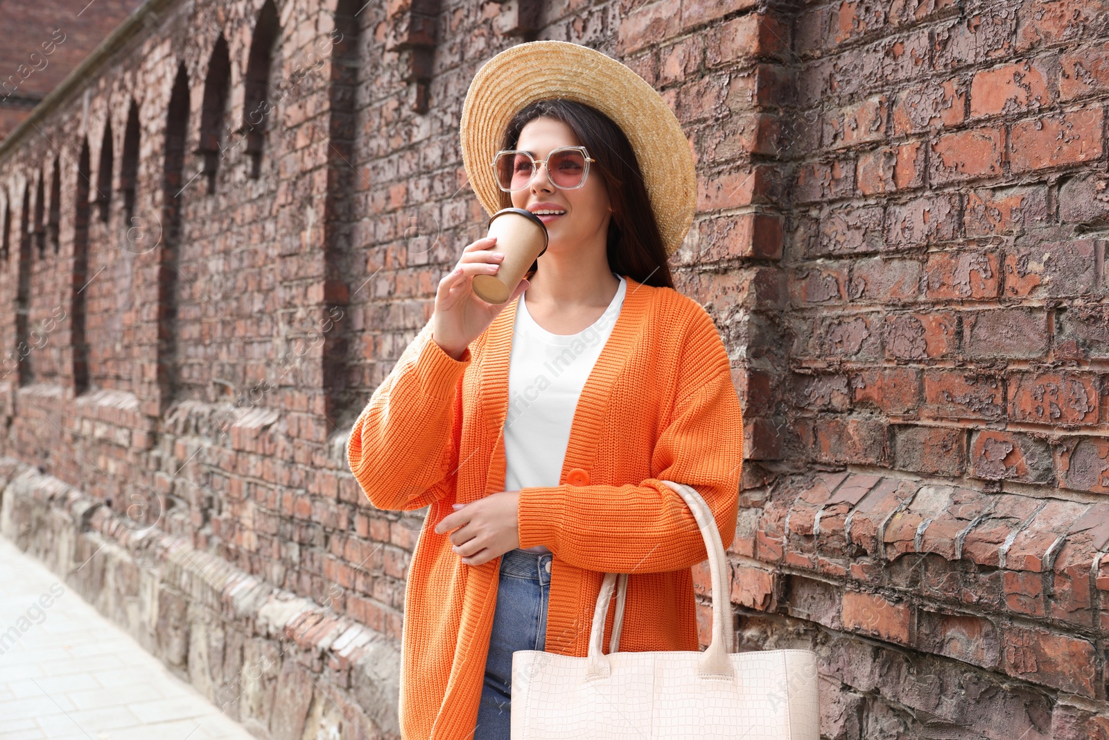 Photo of Young woman with stylish bag and cup of hot drink near red brick wall outdoors