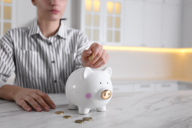 Photo of Woman putting money into piggy bank at white marble table indoors, closeup. Space for text