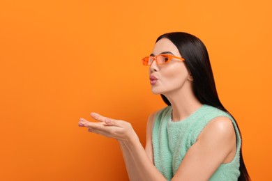 Photo of Beautiful young woman in stylish sunglasses blowing kiss on orange background. Space for text