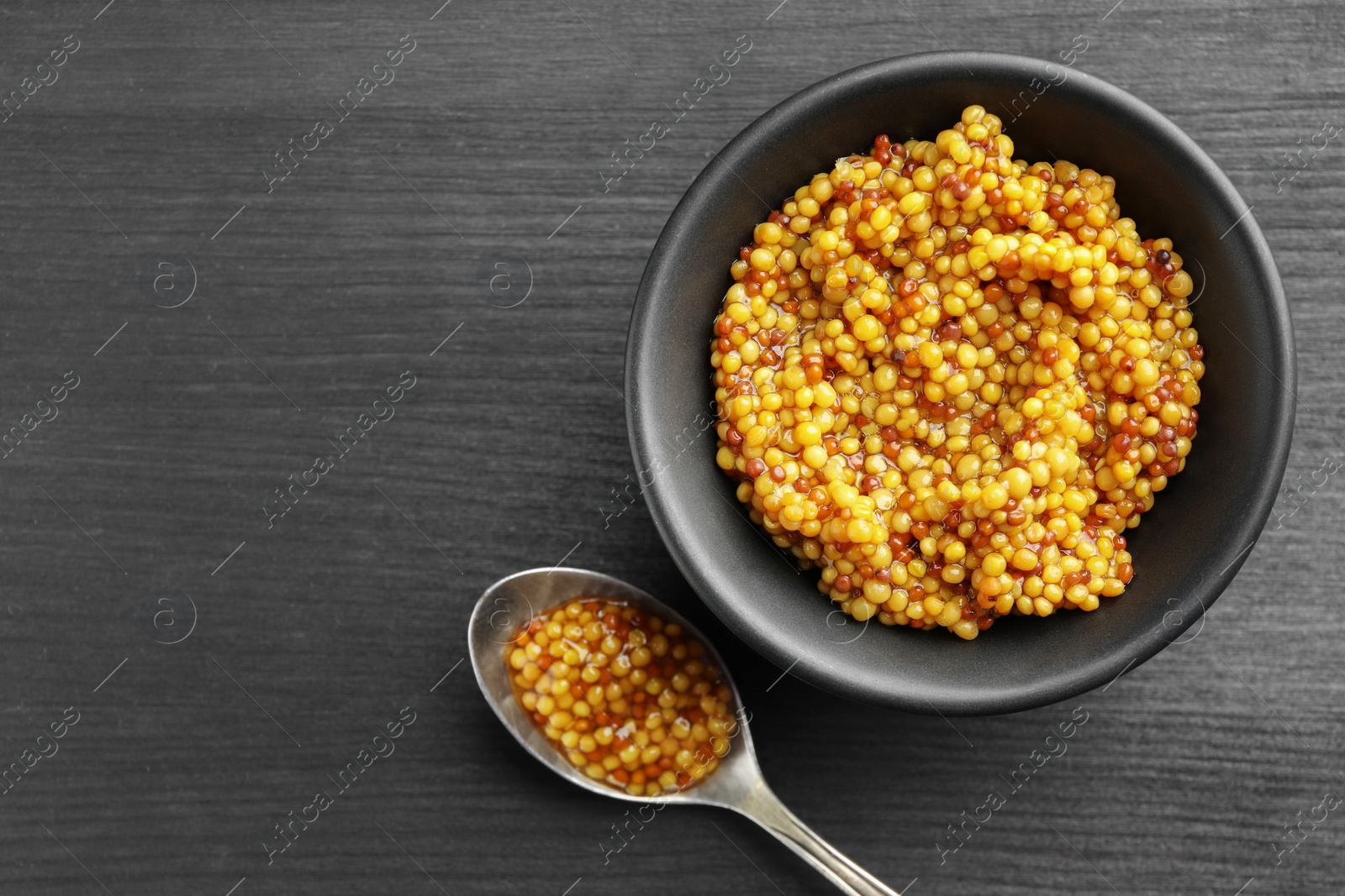 Photo of Bowl and spoon with whole grain mustard on black wooden table, top view. Space for text