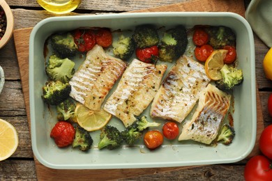 Photo of Pieces of delicious baked cod with vegetables, lemon and spices in dish on wooden table