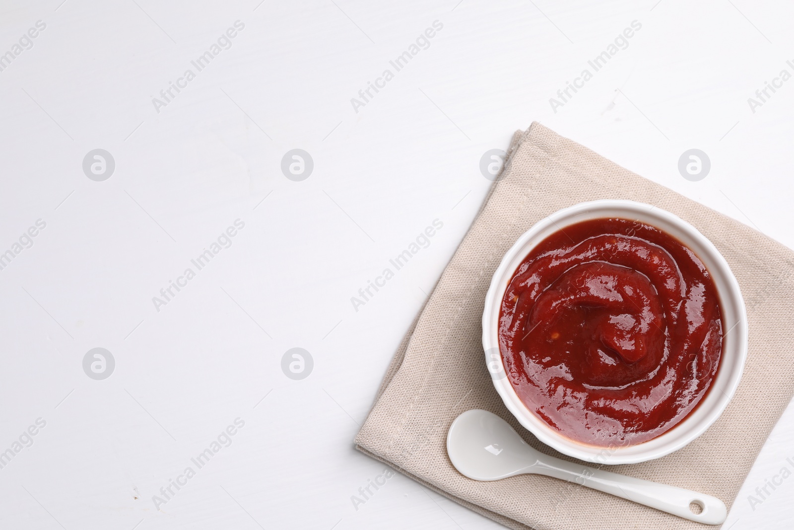 Photo of Organic ketchup in bowl and spoon on white table, top view with space for text. Tomato sauce