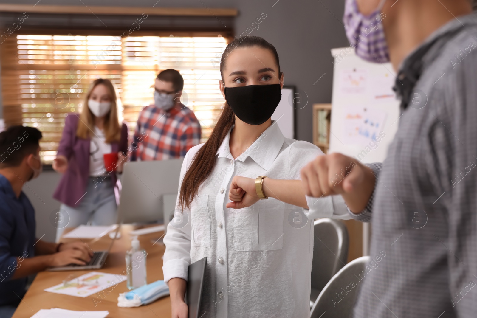 Photo of Coworkers with protective masks making elbow bump in office. Informal greeting during COVID-19 pandemic