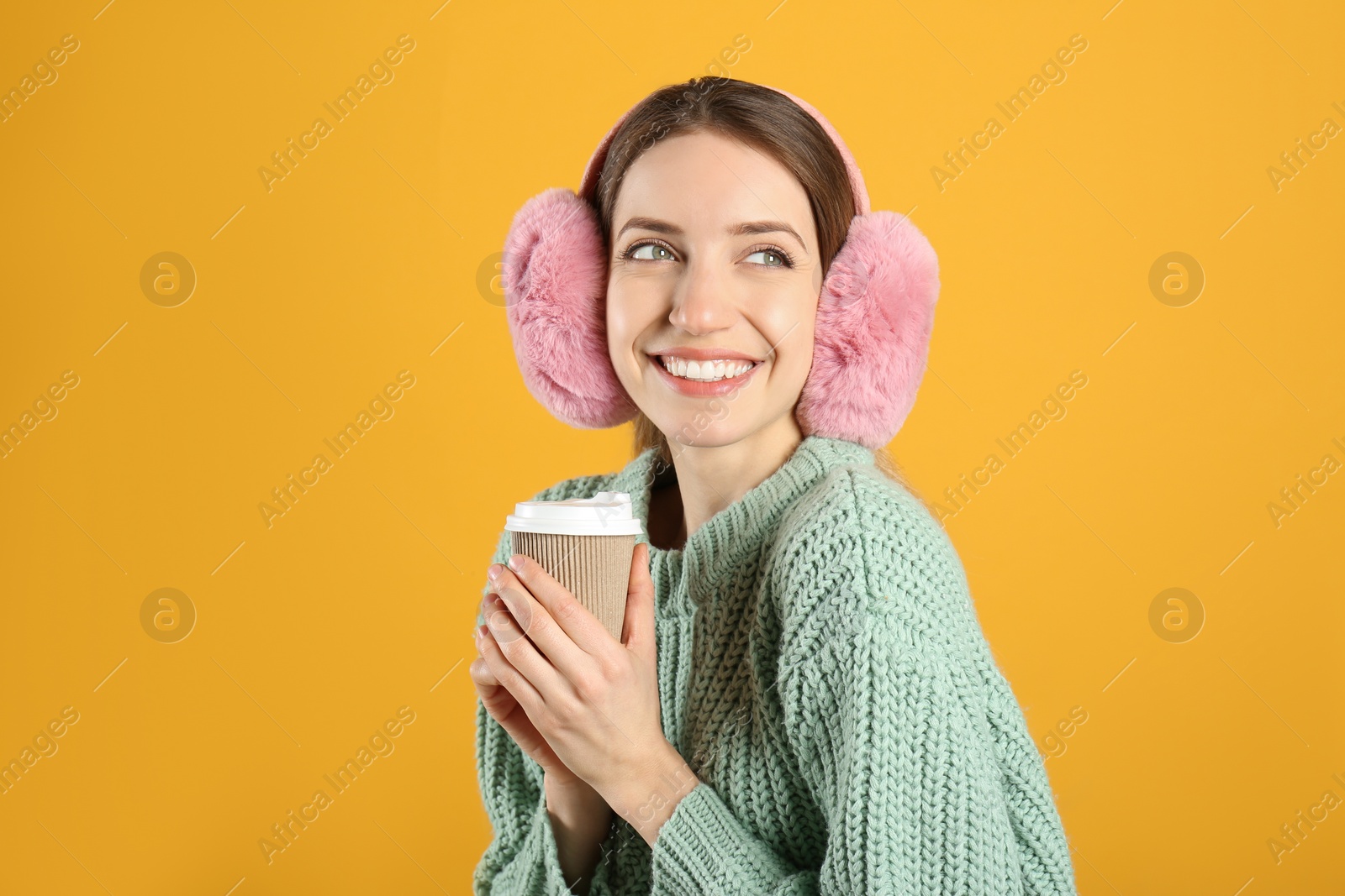 Photo of Happy woman with cup of drink wearing warm earmuffs on yellow background