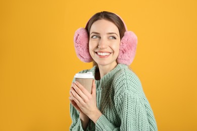 Happy woman with cup of drink wearing warm earmuffs on yellow background