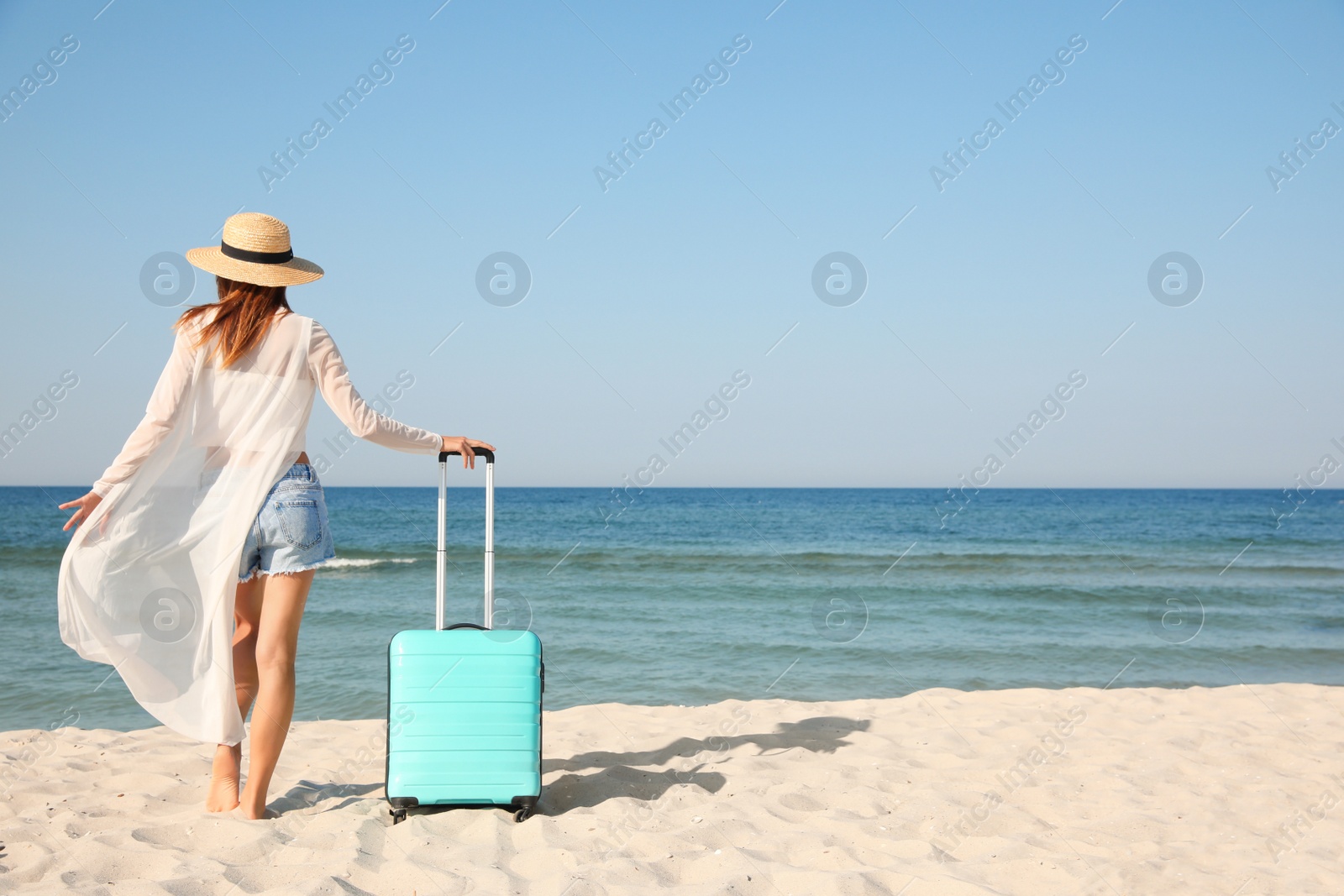 Photo of Woman with suitcase on sandy beach near sea, back view