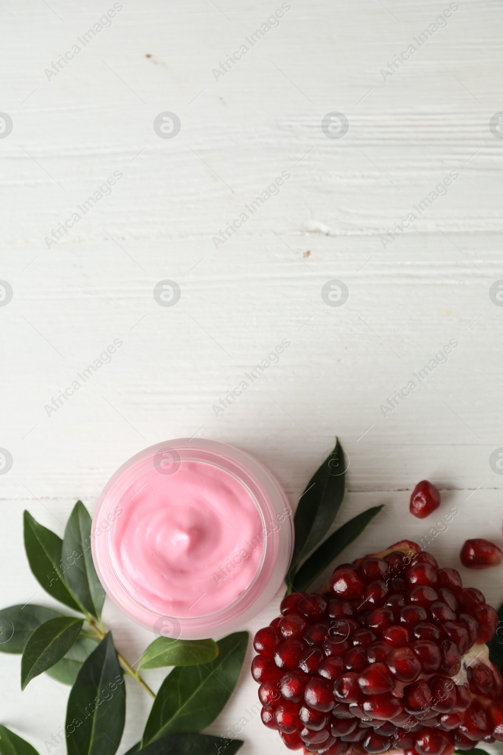 Photo of Jar with natural facial mask, pomegranate seeds and green leaves on white wooden table, flat lay. Space for text