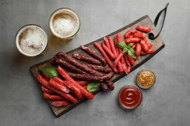 Photo of Different thin dry smoked sausages, sauces and glasses of beer on grey table, flat lay