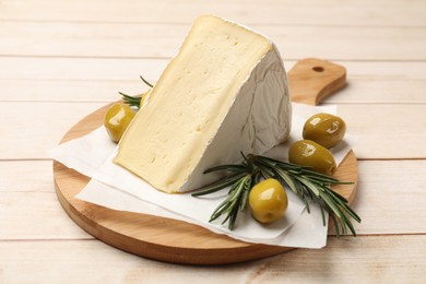 Photo of Board with piece of tasty camembert cheese, olives and rosemary on light wooden table, closeup