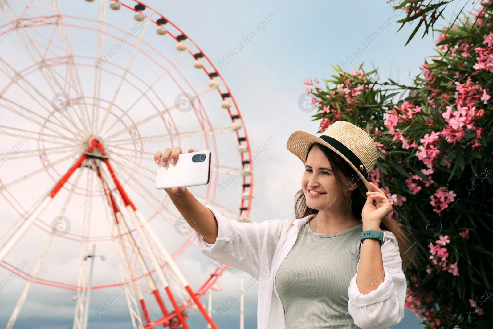 Photo of Woman taking selfie near beautiful blooming oleander and large Ferris wheel outdoors