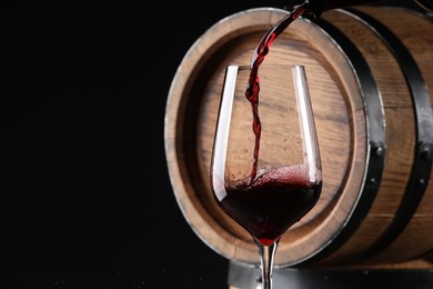 Photo of Pouring red wine into glass near wooden barrel against black background. Space for text