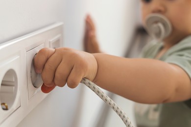 Photo of Cute baby playing with plug and electrical socket at home, closeup. Dangerous situation