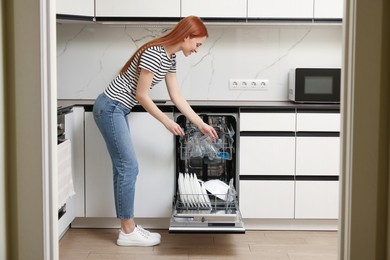 Smiling woman loading dishwasher with glasses and plates in kitchen