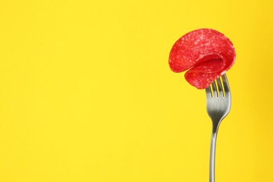 Photo of Fork with tasty slice of salami on yellow background, space for text