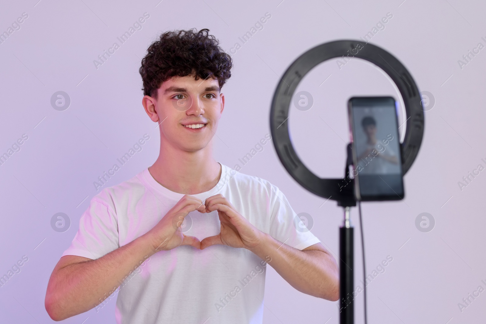 Photo of Smiling teenage blogger making heart gesture to his subscribers while streaming on white background