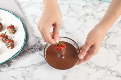 Photo of Woman dipping ripe strawberry into bowl with melted chocolate on table