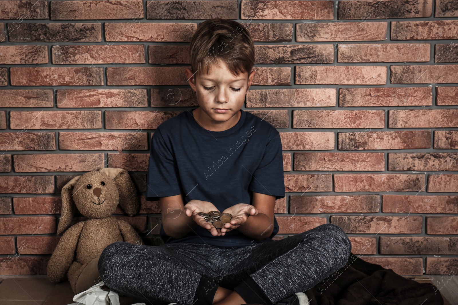 Photo of Poor homeless boy holding coins on floor near brick wall