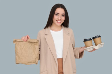 Young female intern holding takeaway cardboard cups and paper bag on grey background