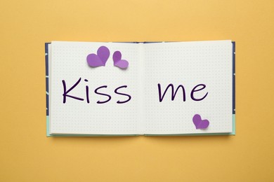 Open notebook with phrase Kiss Me and paper hearts on pale orange background, top view