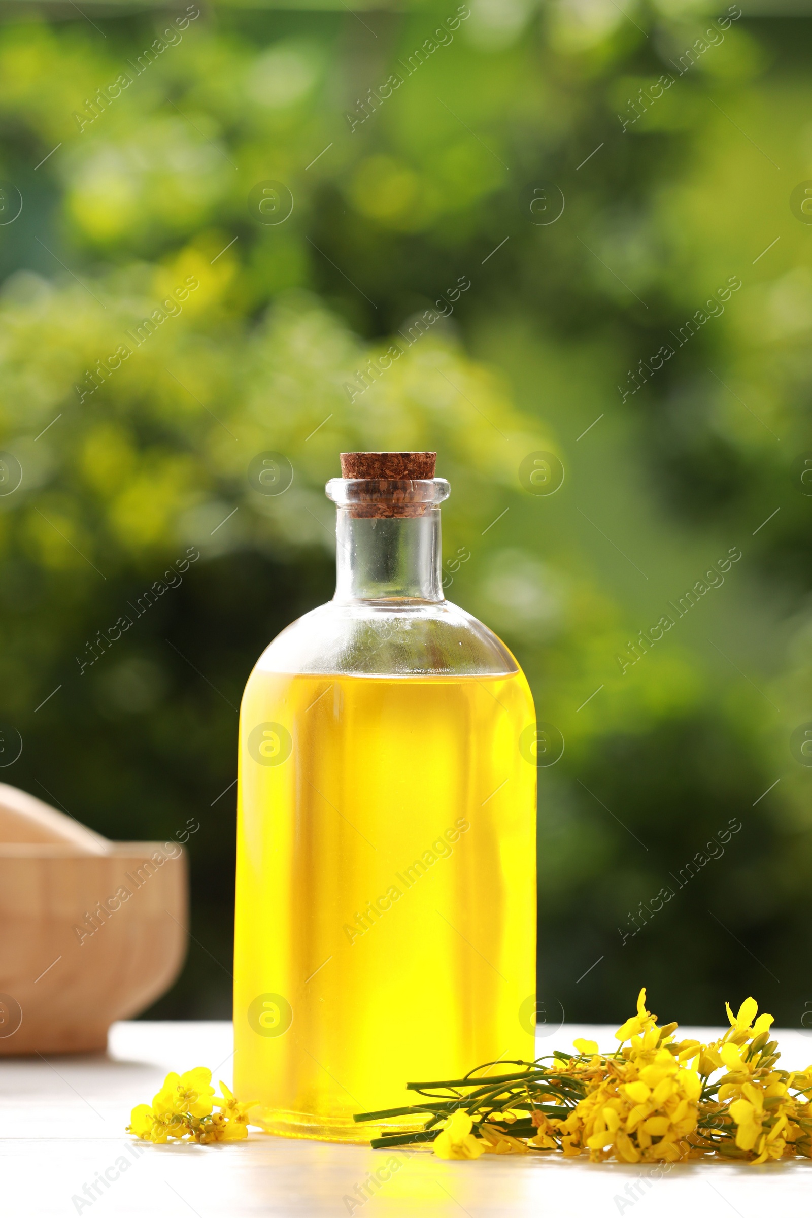 Photo of Rapeseed oil in glass bottle and yellow flowers on white wooden table outdoors