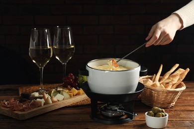 Photo of Woman dipping piece of bread into fondue pot with melted cheese at wooden table, closeup