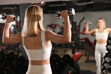 Woman training with dumbbells near mirror in gym, back view