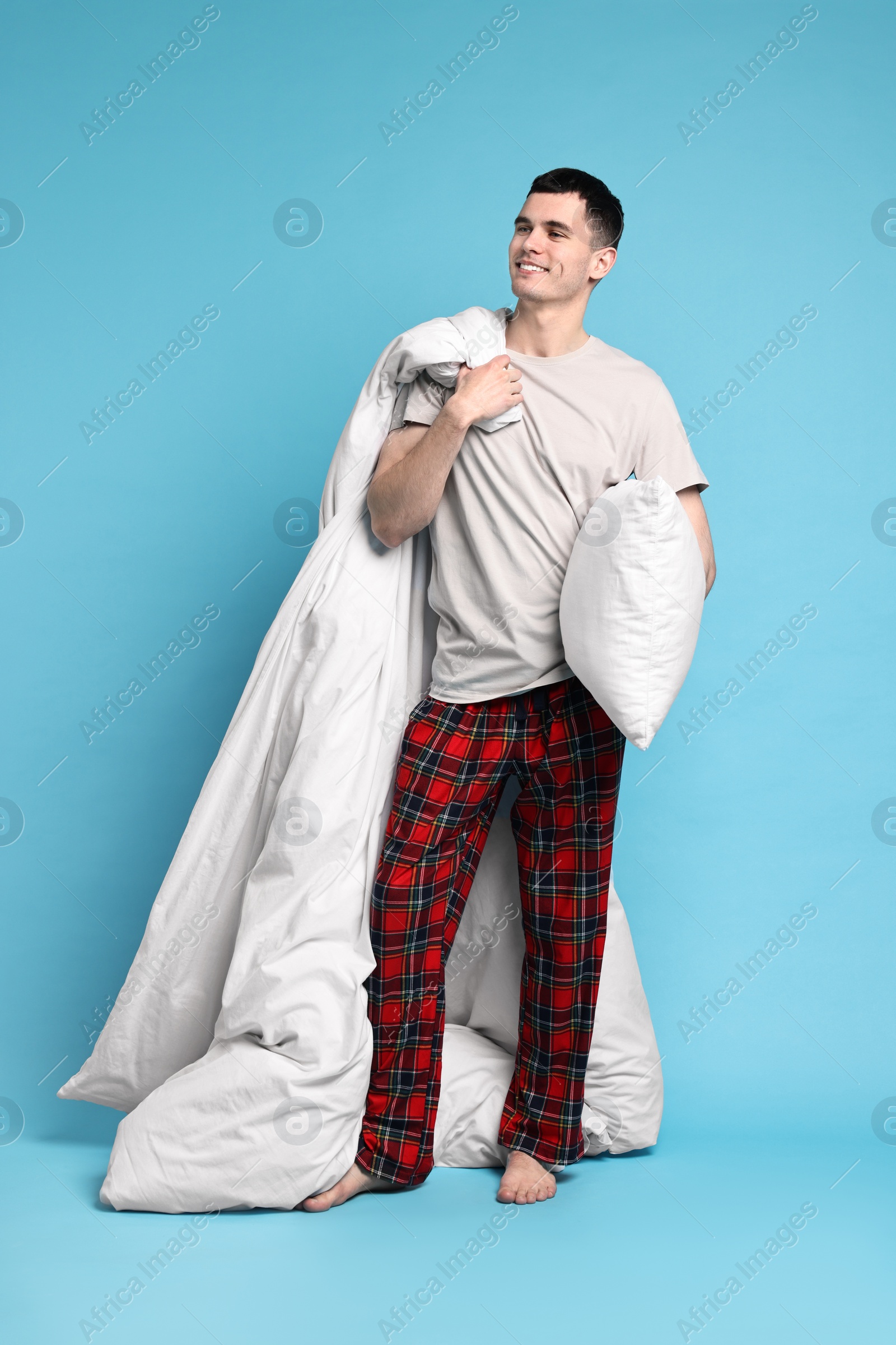 Photo of Happy man in pyjama holding blanket and pillow on light blue background