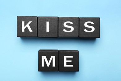 Black wooden cubes with phrase Kiss Me on light blue, flat lay
