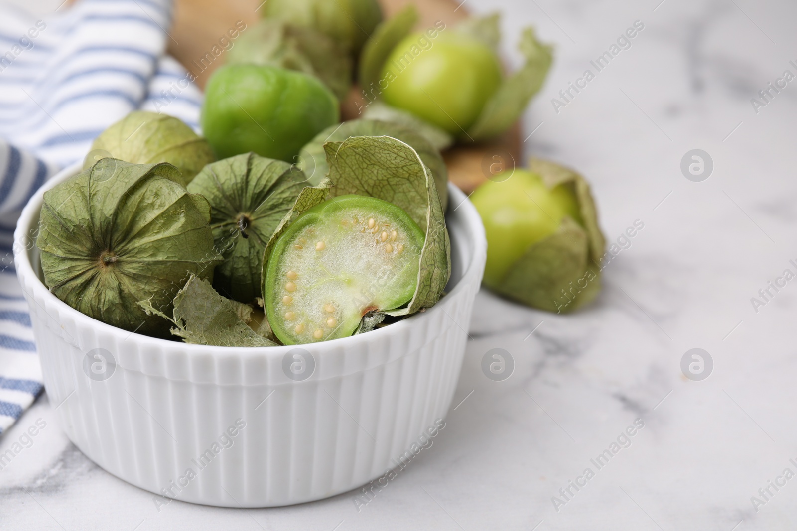 Photo of Fresh green tomatillos with husk in bowl on light marble table, closeup