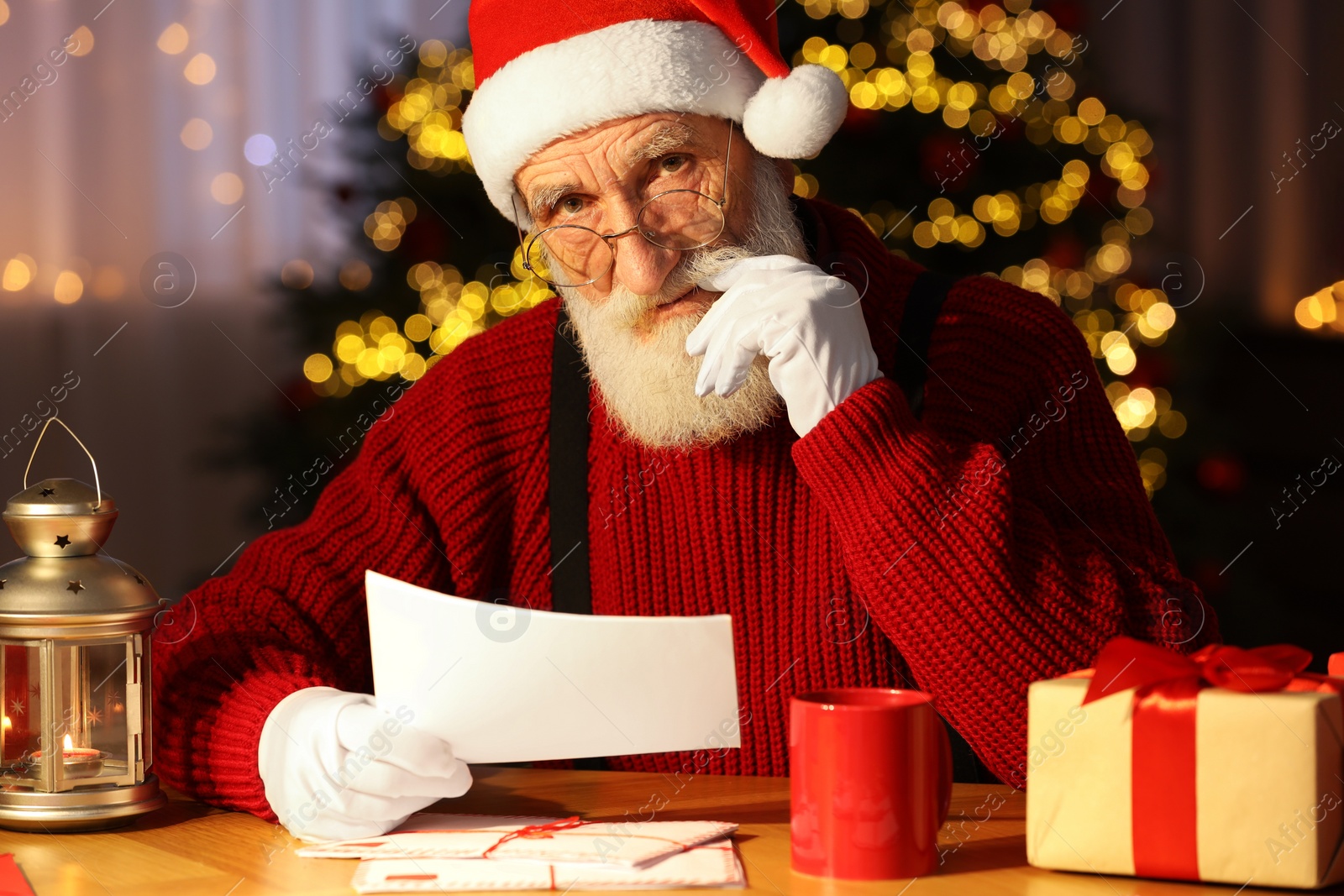 Photo of Santa Claus reading letter at his workplace in room with Christmas tree