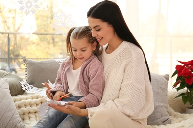 Photo of Mother and daughter making paper snowflake near window at home