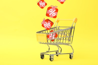 Image of Discount offer. Gift boxes with percent signs falling into shopping trolley on yellow background, space for text