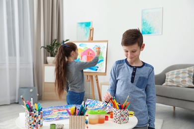Photo of Boy painting at table and girl near easel indoors. Creative children