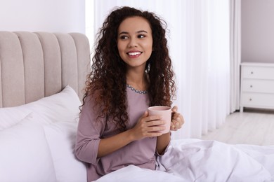 Happy African American woman with cup of drink in bed at home