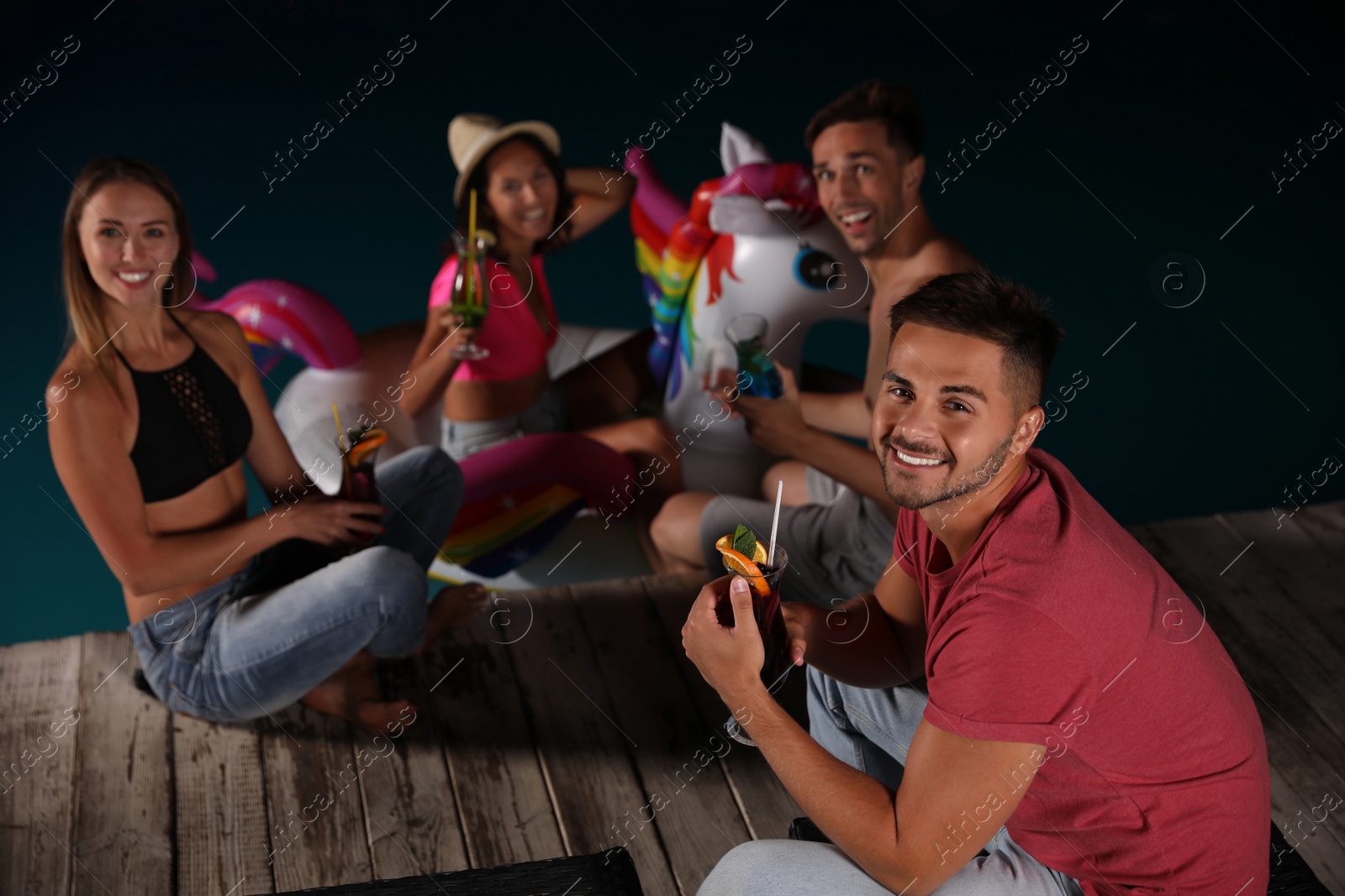 Photo of Happy young friends with refreshing cocktails relaxing near outdoor swimming pool at night