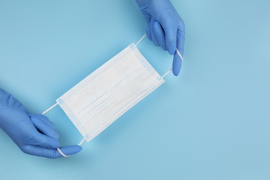 Photo of Doctor in medical gloves holding protective mask on light blue background, closeup. Space for text