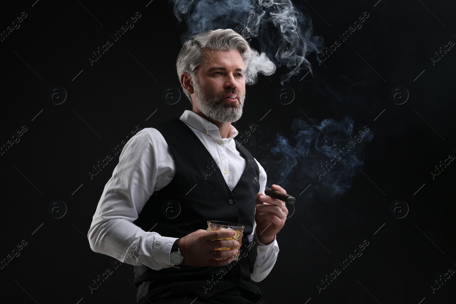 Photo of Bearded man with glass of whiskey smoking cigar against black background