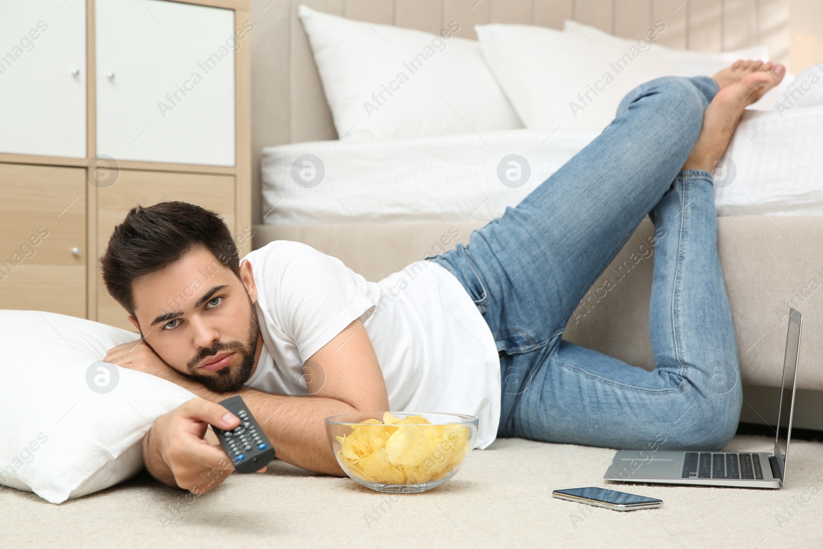 Photo of Lazy young man with bowl of chips watching TV while lying on floor at home