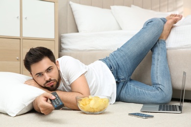 Photo of Lazy young man with bowl of chips watching TV while lying on floor at home