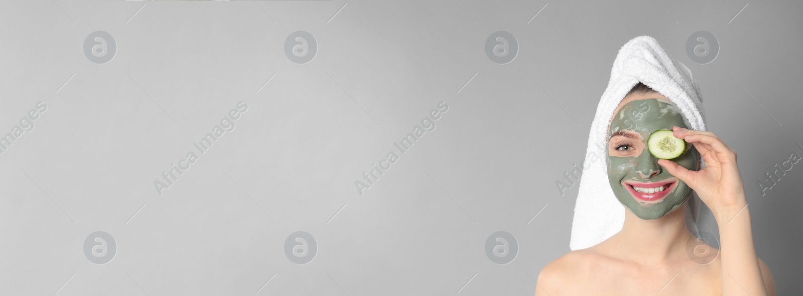 Image of Young woman with clay mask on her face holding cucumber slice against grey background, space for text. Banner design