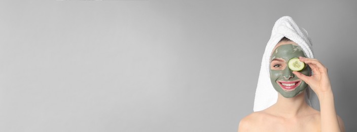 Image of Young woman with clay mask on her face holding cucumber slice against grey background, space for text. Banner design