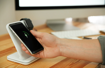 Photo of Man taking mobile phone from wireless charger at wooden table, closeup. Modern workplace accessory