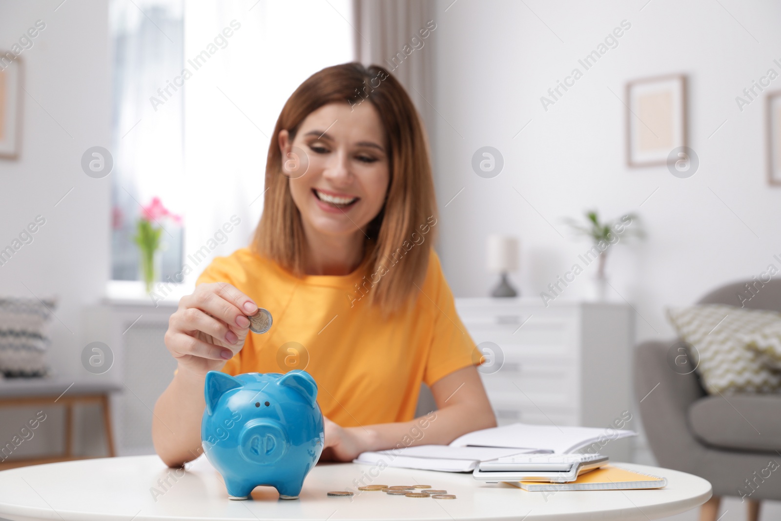 Photo of Woman putting coin into piggy bank at table in living room. Saving money