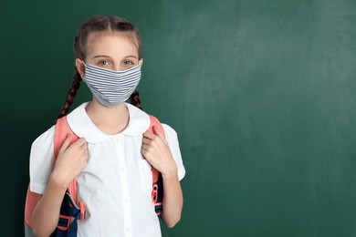 Schoolgirl wearing protective mask near green chalkboard, space for text. Child's safety from virus