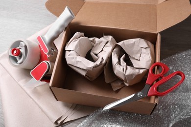 Open box with wrapped items, adhesive tape, scissors, paper and bubble wrap on wooden table