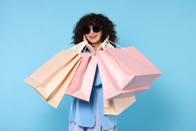 Happy young woman with shopping bags and stylish sunglasses on light blue background