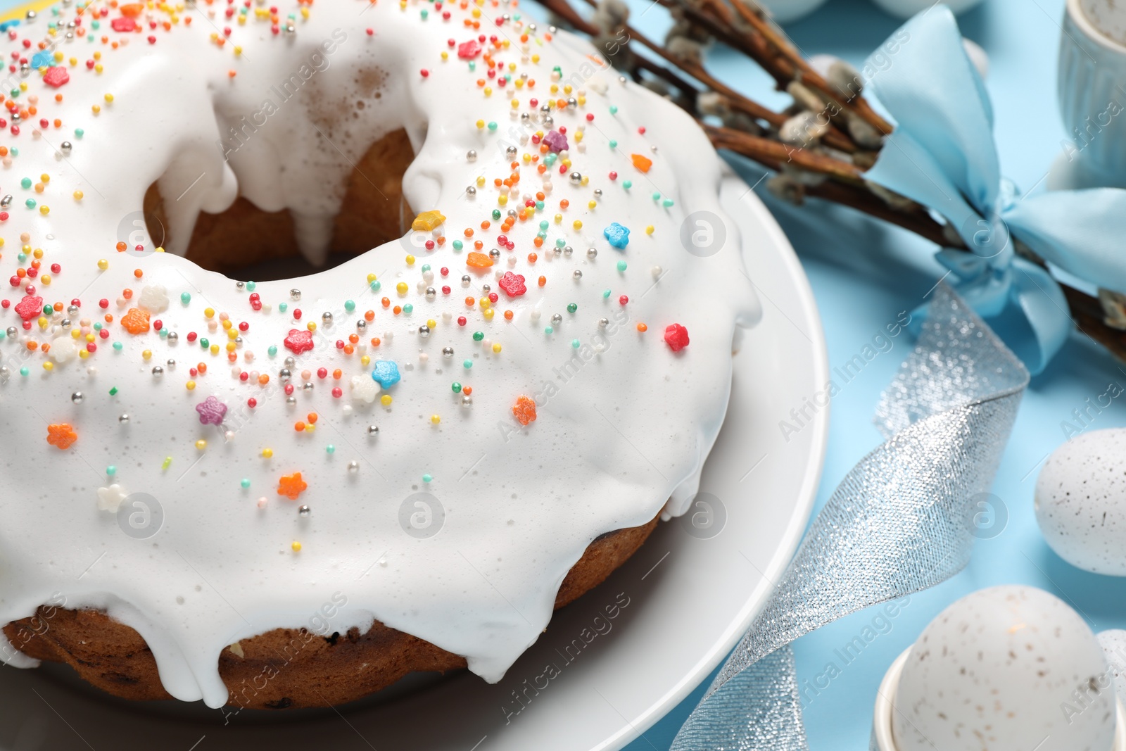 Photo of Delicious Easter cake decorated with sprinkles near painted eggs and willow branches on light blue background, closeup