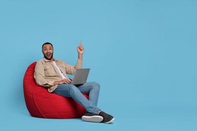 Smiling young man with laptop sitting on beanbag chair against light blue background