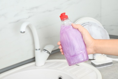 Photo of Woman holding bottle of cleaning product for dish washing near kitchen sink, closeup