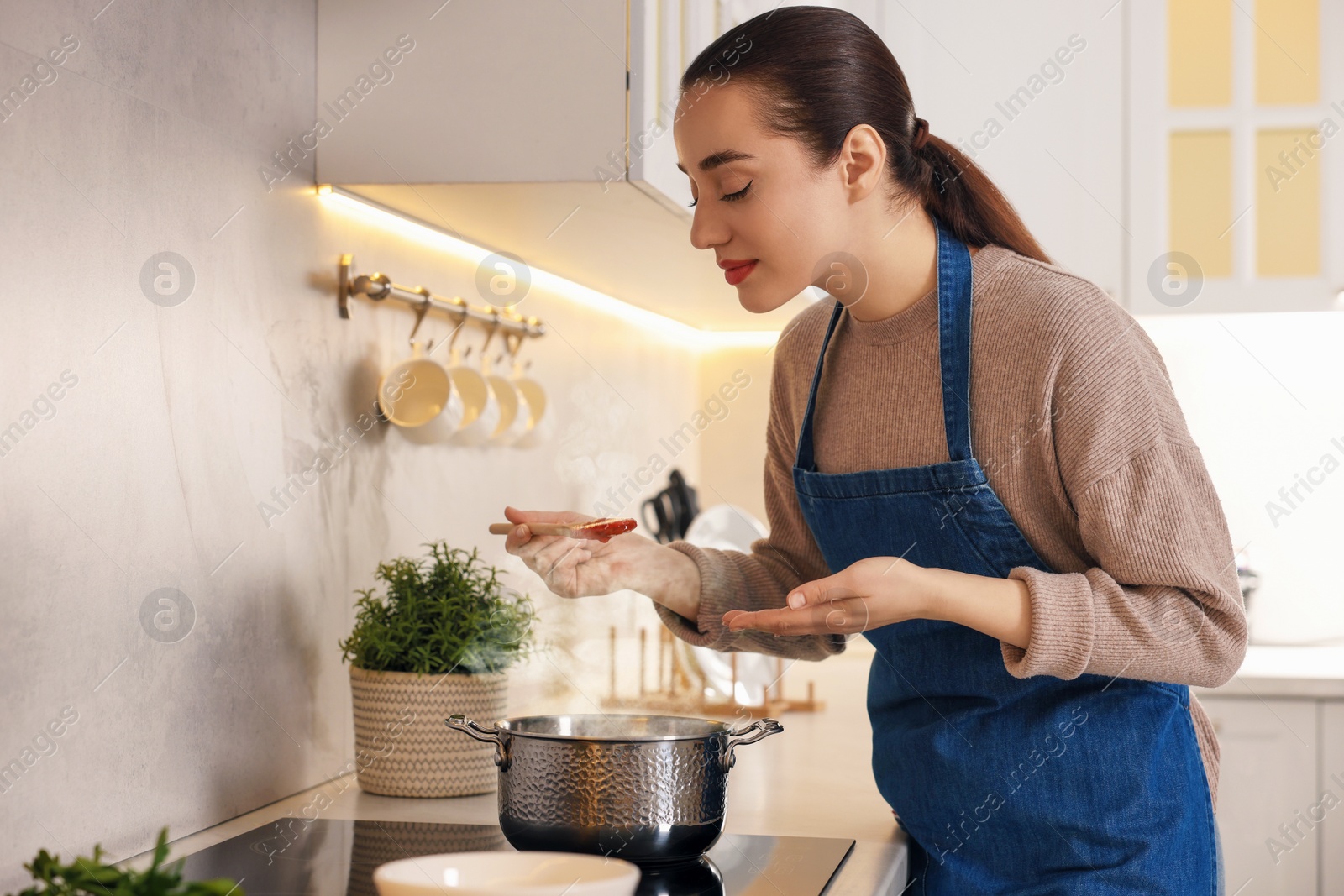Photo of Smiling woman with wooden spoon tasting tomato soup in kitchen. Space for text
